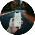 Ein Smartphone, auf dem eine Navigations-App geöffnet ist. Im Hintergrund eine Straße mit herbstlichen Laub.