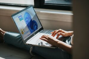 Ein eingeschalteter Laptop auf dem Schoß einer Frau vor einem Fenster.